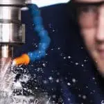 Close up image of a man wearing goggles watching a drill piece and coolant drilling into metal