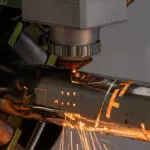 A close-up of a Tube Laser cutting out intricate shapes on a stainless steel tube. Sparks are flying out of the tube, and you can see the chuck of the machine in the background holding the non-cut end of the tube.