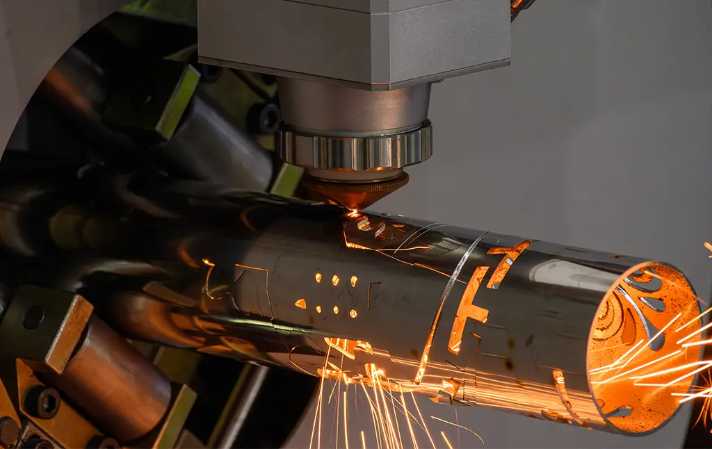 A close-up of a Tube Laser cutting out intricate shapes on a stainless steel tube. Sparks are flying out of the tube, and you can see the chuck of the machine in the background holding the non-cut end of the tube.